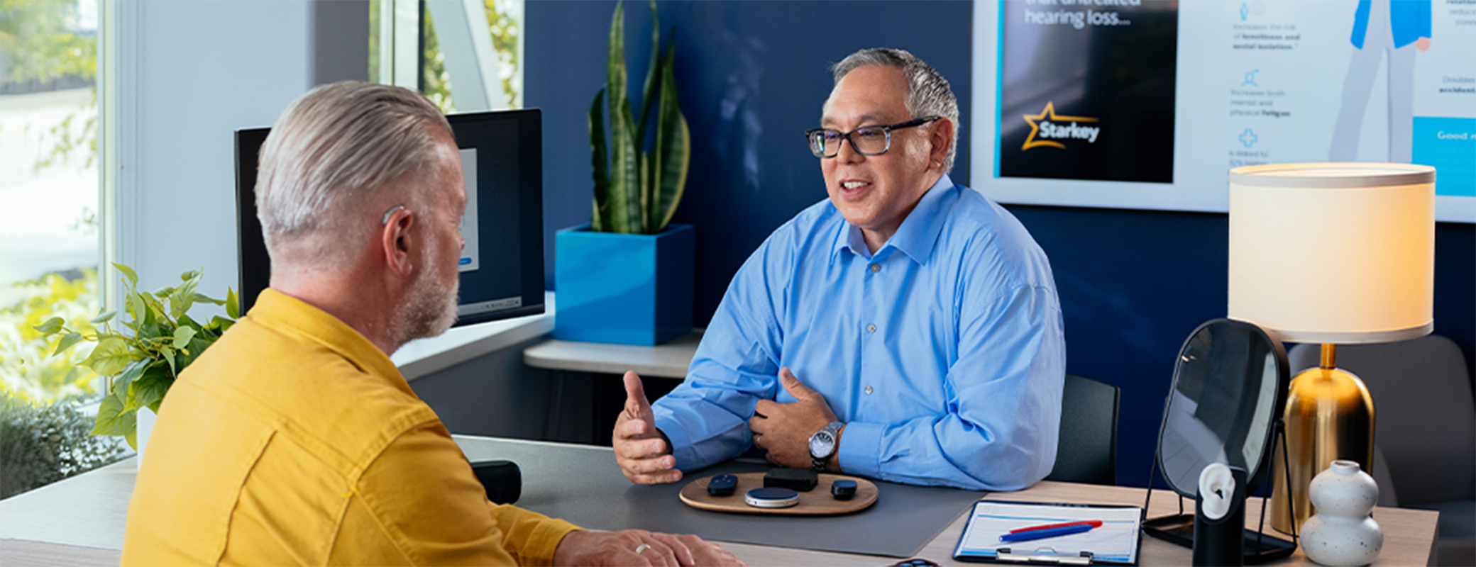 Image of a male hearing care professional talking to a male patient from his desk